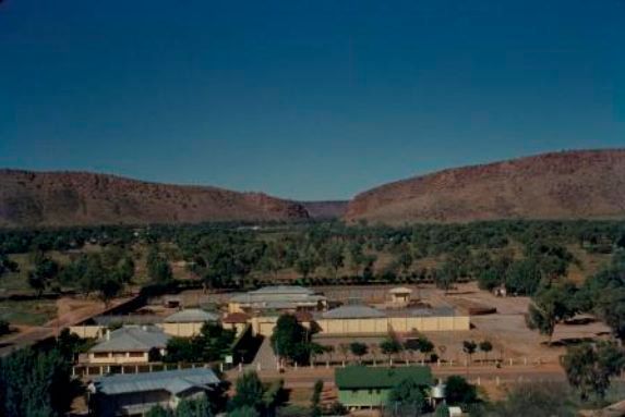A desert village with low buildings and trees beneath red sandstone cliffs under a clear blue sky