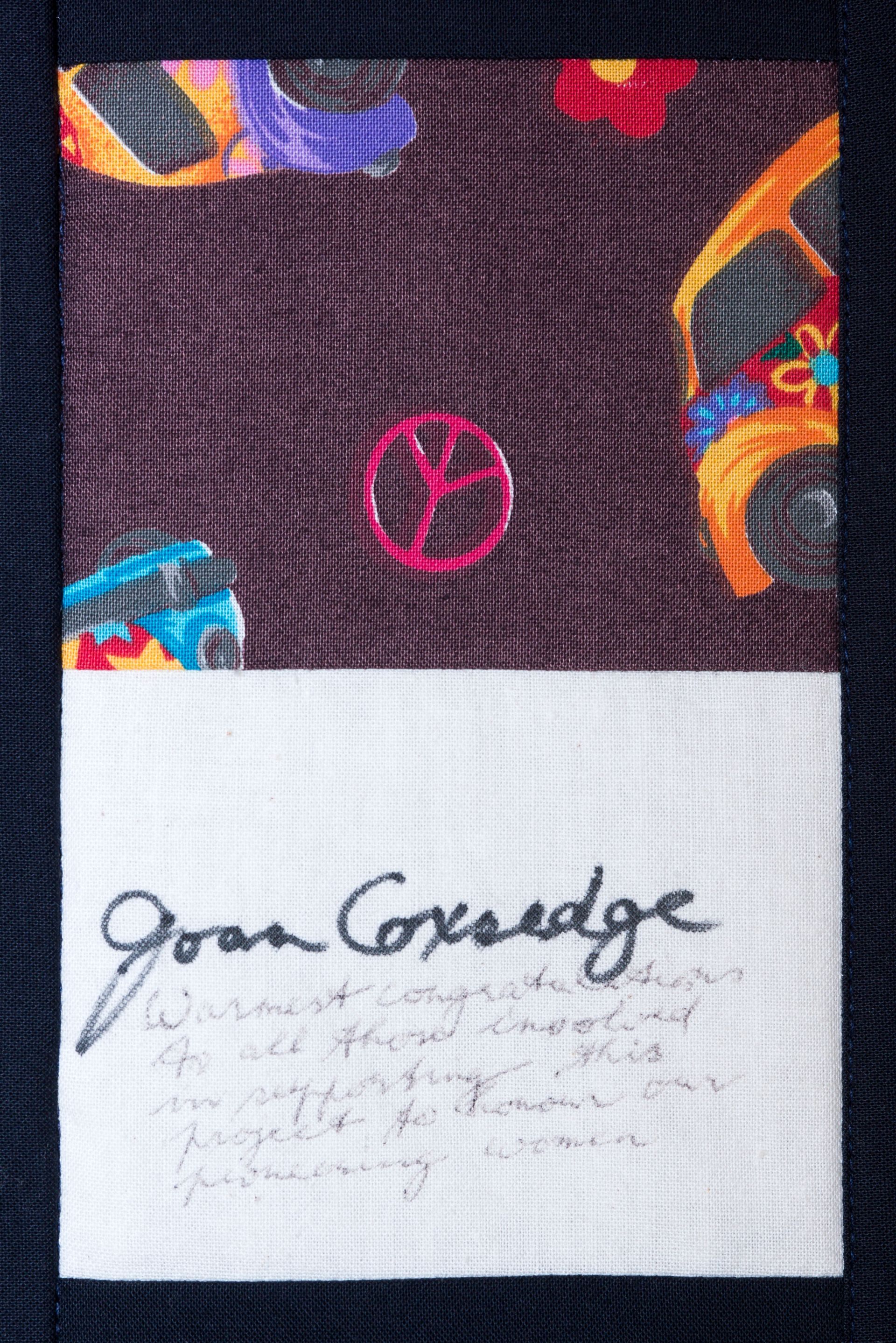 Close-up of a dark patterned fabric with a white label signed “Joan Crouch” and faint handwritten notes.