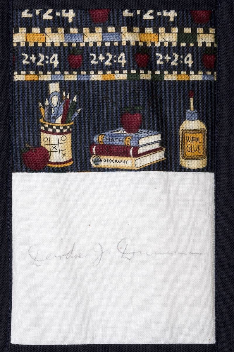 A small embroidered wall hanging with French phrases, wine bottle, books, and white blank rectangle on dark fabric.