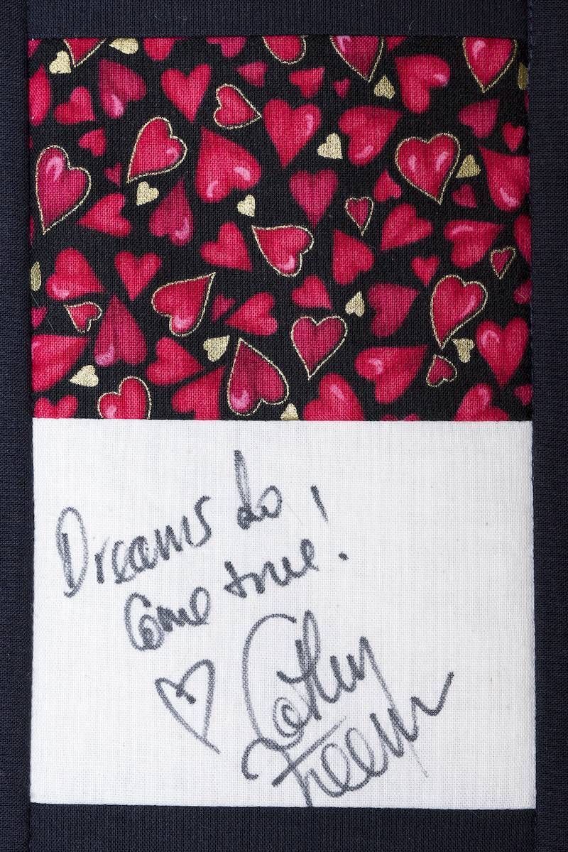 Handwritten note on white paper over red heart-patterned fabric, reading “Dreams do come true” with a heart and signature.