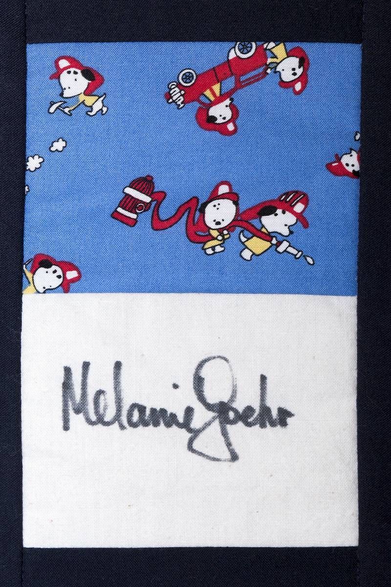 Blue patterned tie with red cartoon figures and a white label reading “Melanie Seher”