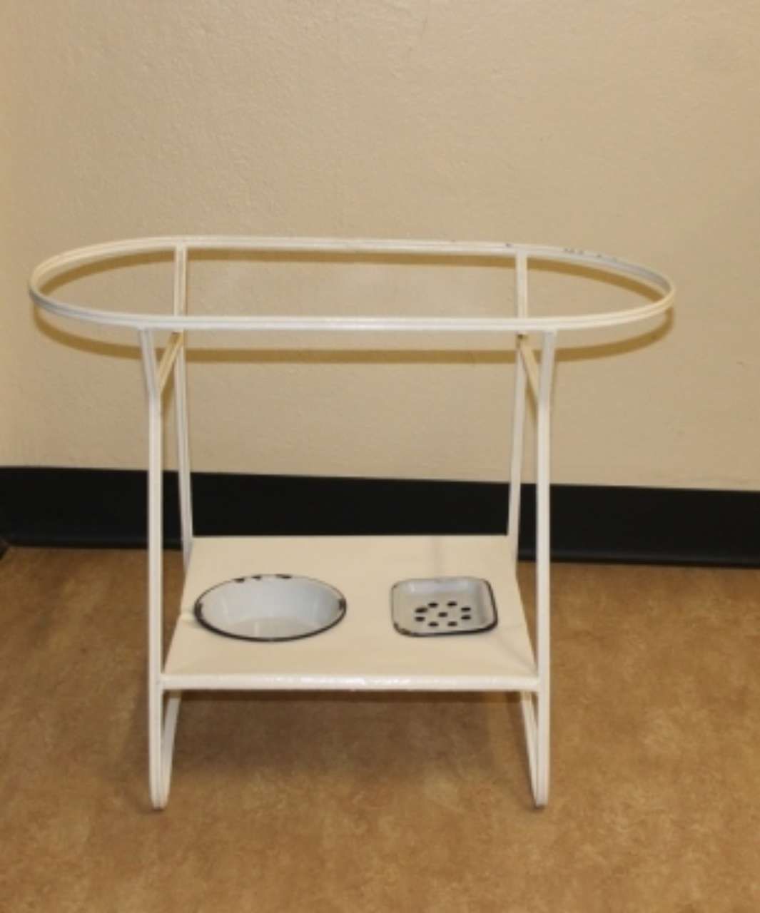 White metal pet feeding stand with oval tray on a beige floor against a wall.