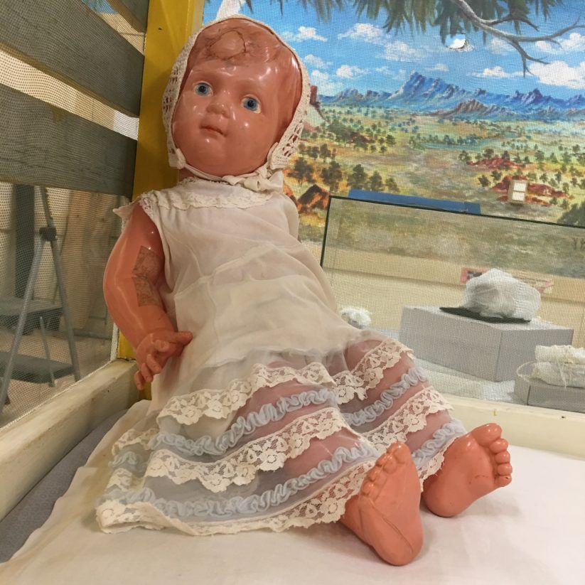 Porcelain doll in a white lace dress sitting on a bench with a painted landscape backdrop