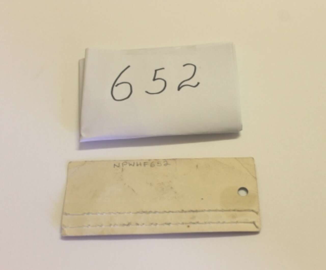 White folded note labeled “652” above a beige tag with a hole on a plain wall.