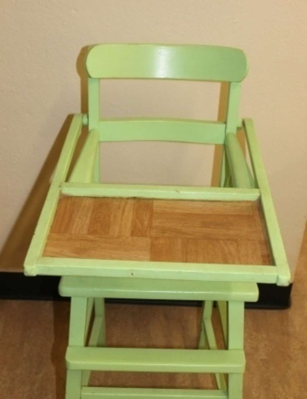 Light green wooden high chair with tray against a beige wall