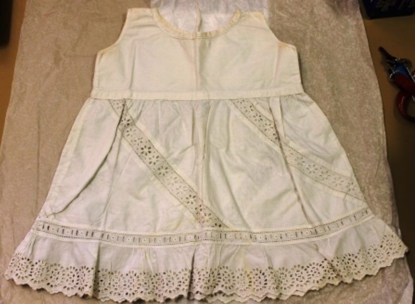 White sleeveless lace dress with ribbon detailing, laid flat on a beige surface.