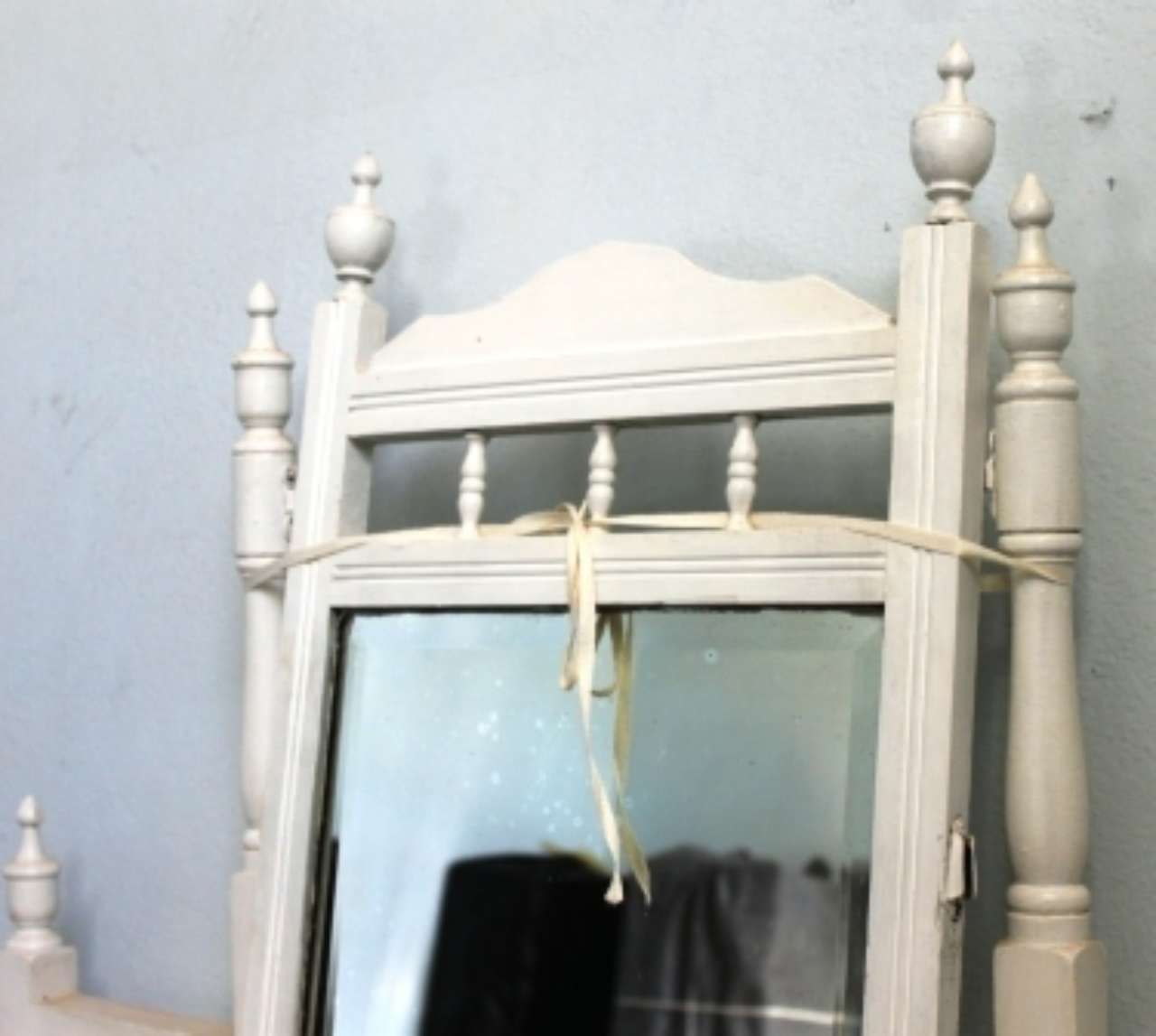 White ornate mirror with a ribbon tied across the top, leaning against a light blue wall.