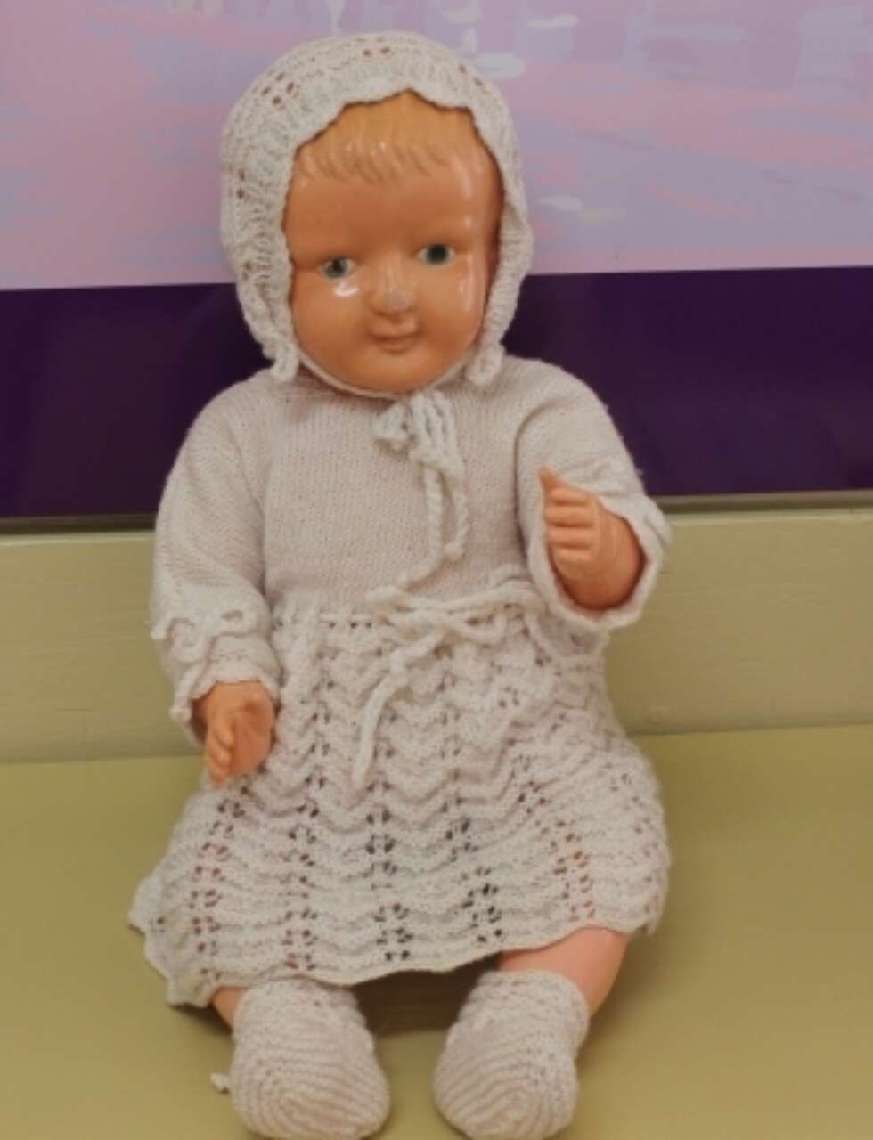 Porcelain doll in a white crocheted dress and bonnet, seated against a purple background.