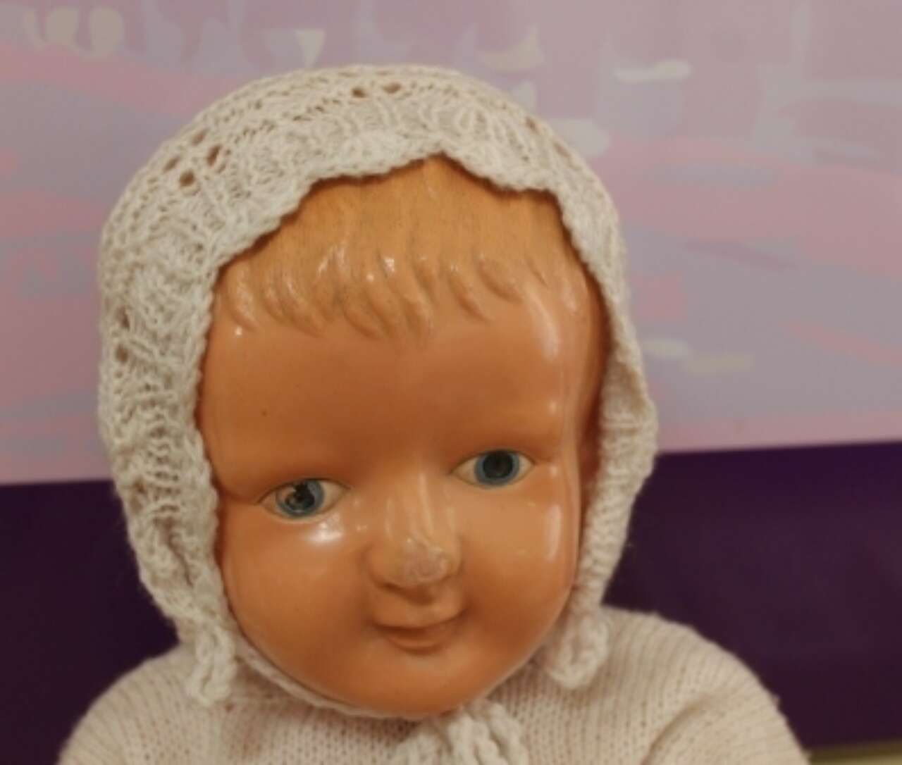 Cream-colored doll head with a knit bonnet, shown against a purple background