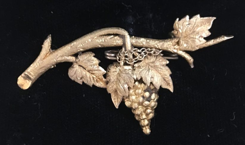 Gold grapevine-shaped brooch with leaves and a hanging cluster of grapes on a black background