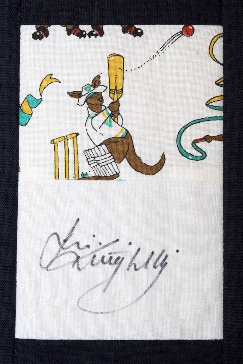 Illustrated cricket scene on white fabric with a handwritten signature below.
