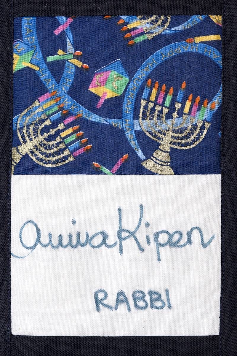 Embroidered blue fabric with abstract shapes and the words “Qui a kiper” and “RABBI”