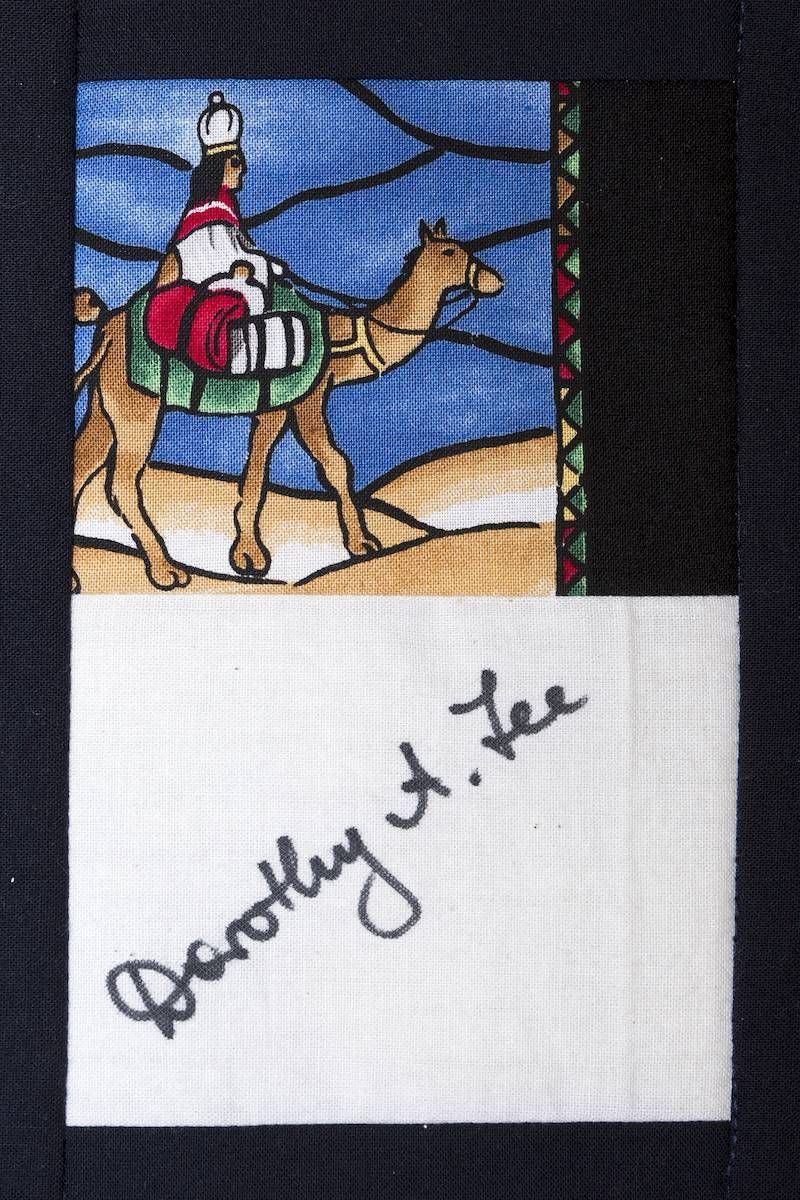 Camel and rider in colorful mosaic above a handwritten signature on white fabric.