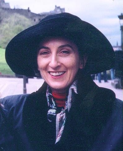 Smiling person in a wide black hat and dark coat outdoors near buildings and trees