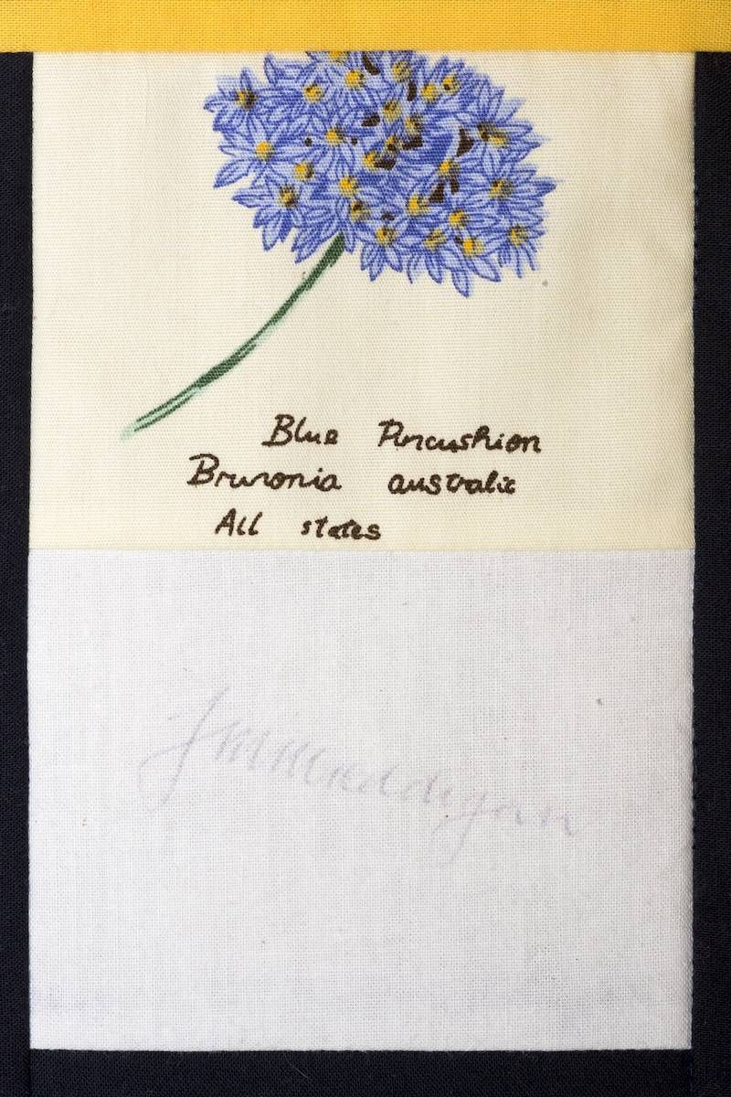 Blue thistle flower embroidered on white cloth with handwritten label beneath it