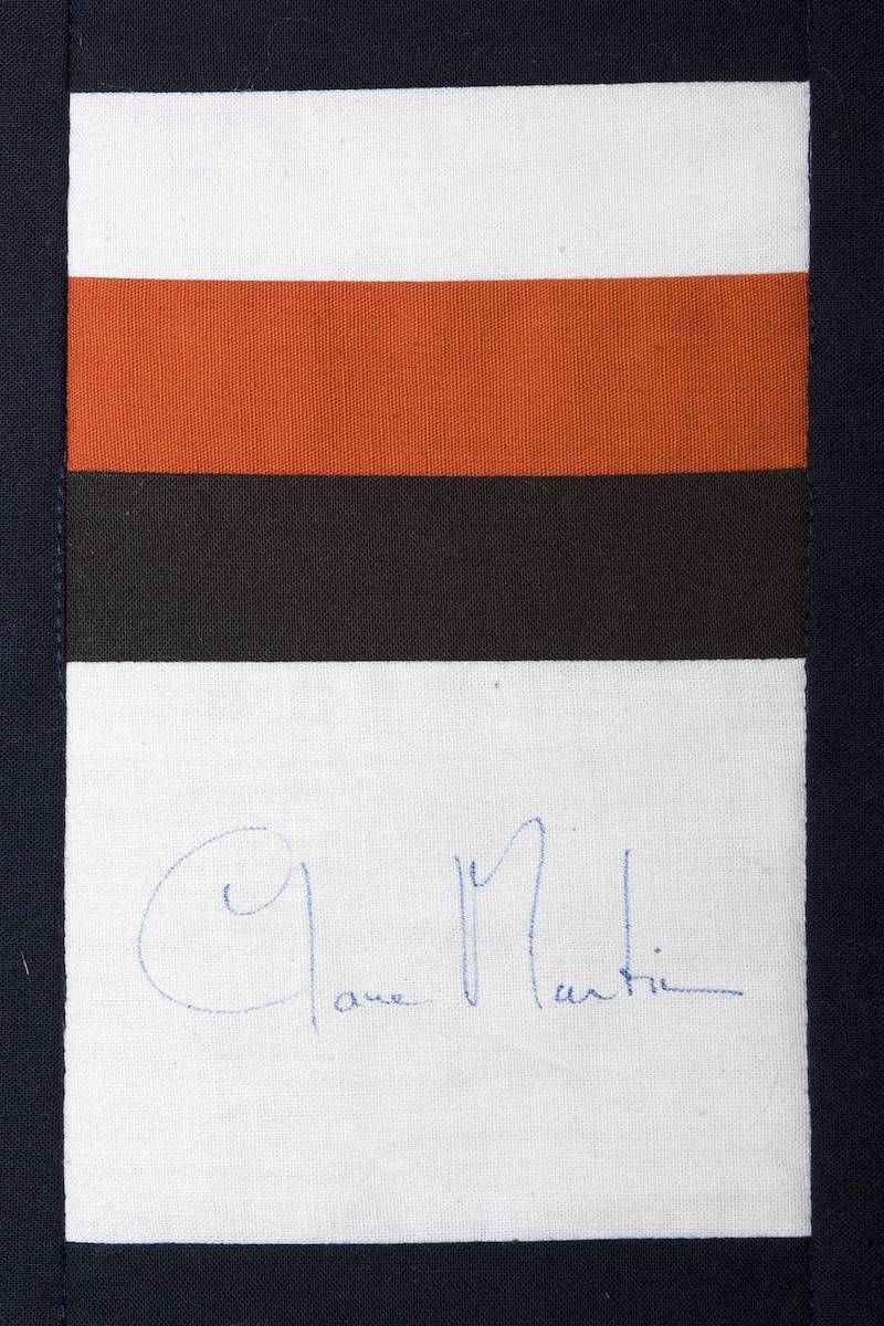 Close-up of a fabric patch with white, orange, and black stripes above a faint handwritten signature.