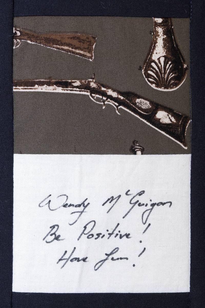 Handwritten note beneath a small metallic object on dark patterned fabric.