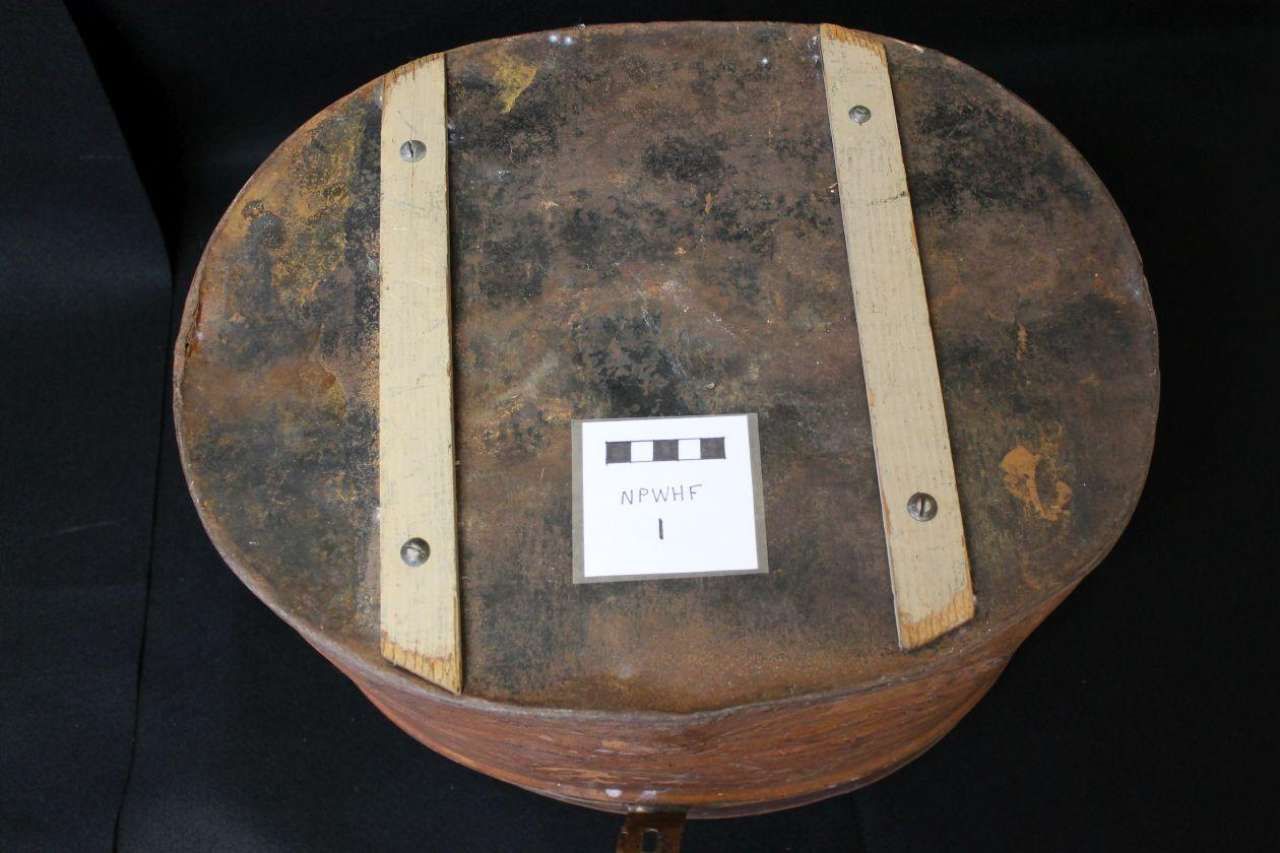 Round brown wooden box lid with two beige straps and a small white label centered on top