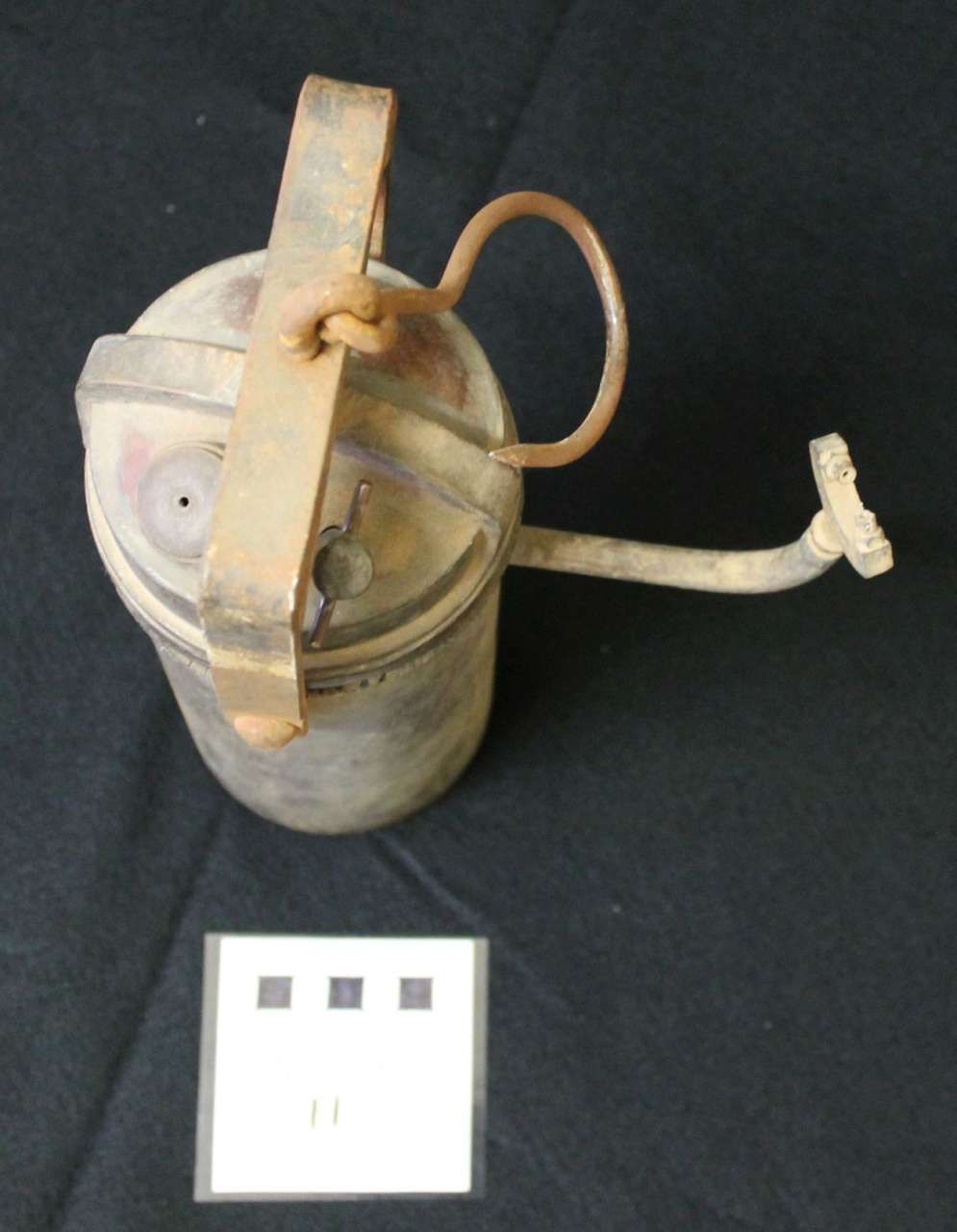 Old metal canister with a handle and looped hose on a dark background, beside a small label card.