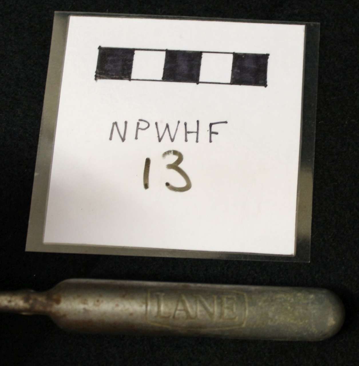 White evidence card labeled “NPWHF 13” above a metal cylindrical object on a dark background