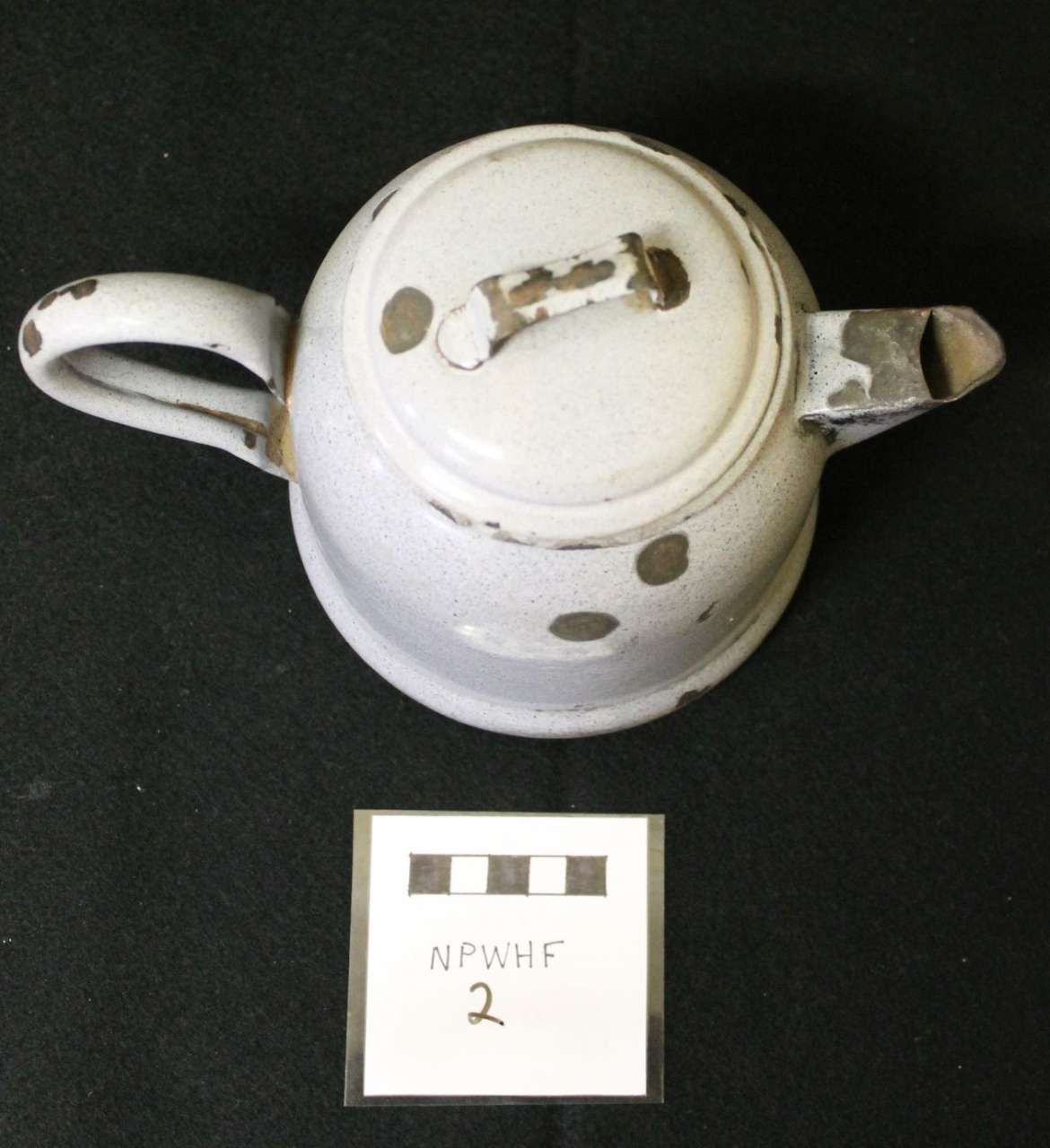 White ceramic teapot with lid and handle, shown on a black background beside a small label.