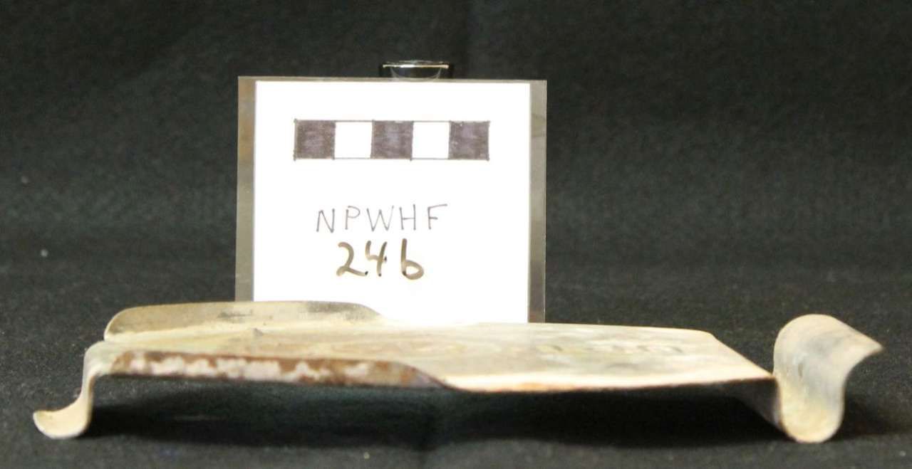 Small beige metal object with curled ends, displayed in front of a labeled box on a dark background