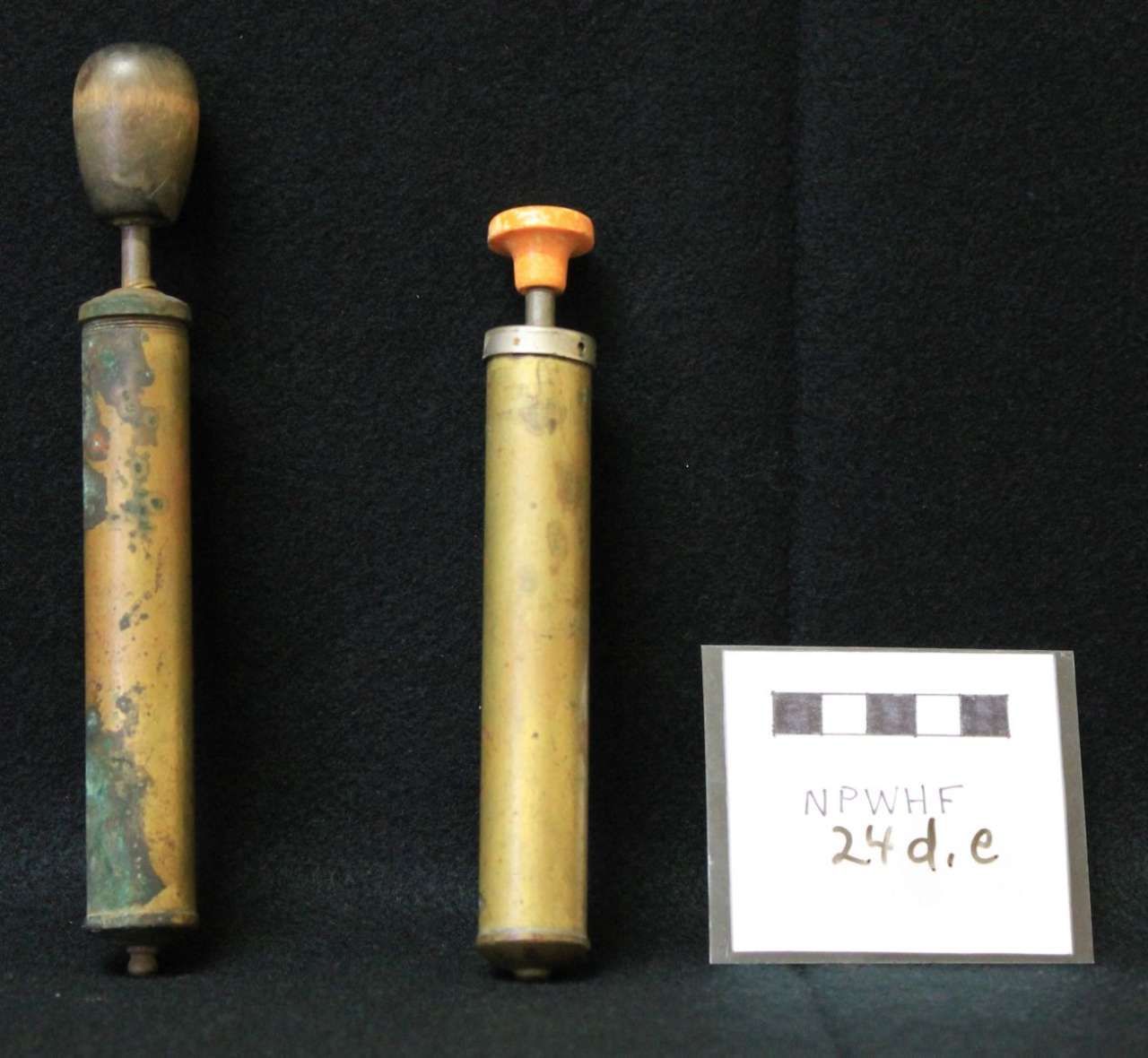 Two old metal cylindrical tools, one with a wooden knob and one with an orange knob, beside a small label card.