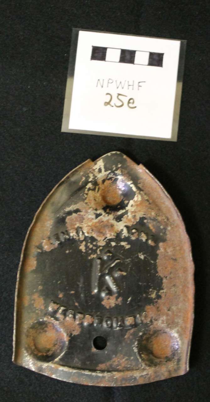 Rusty metal bracket or plate with two bolts and a hole, beneath a small handwritten label.