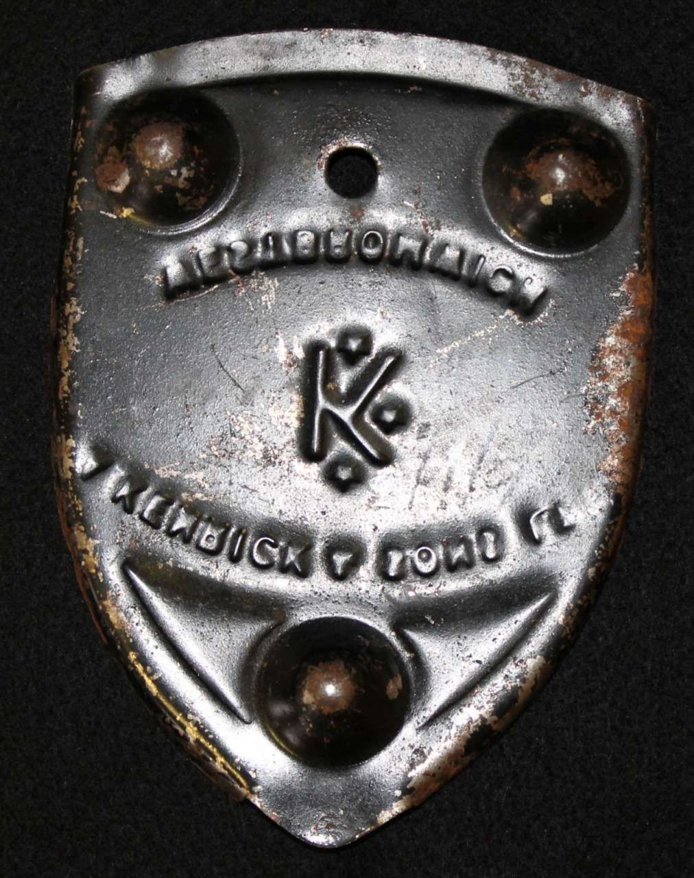 Metal shield-shaped badge with embossed logo and text, held by three rivets on a dark background