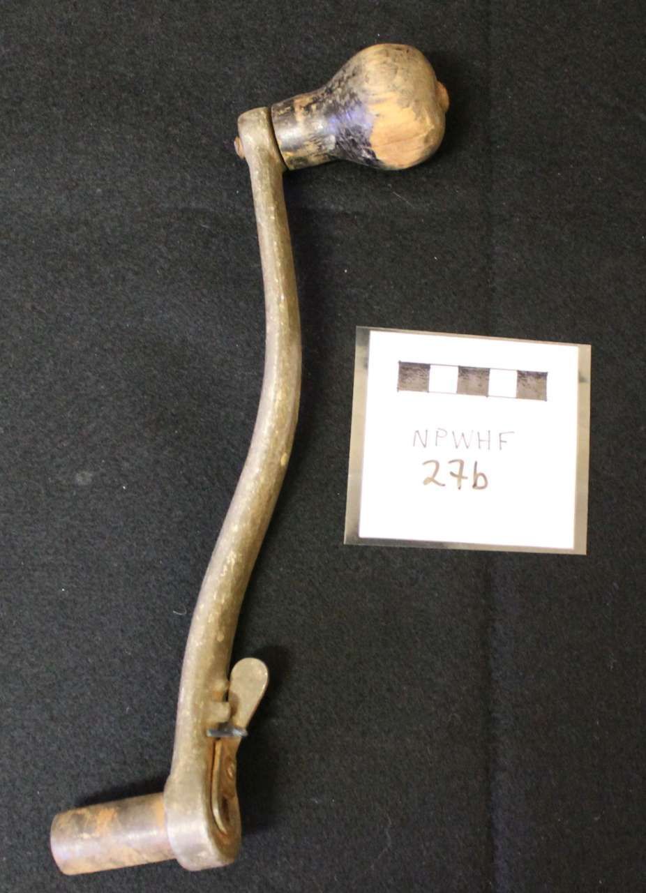 Curved metal hand crank with wooden knob and cylindrical socket, beside a small labeled tag on black background