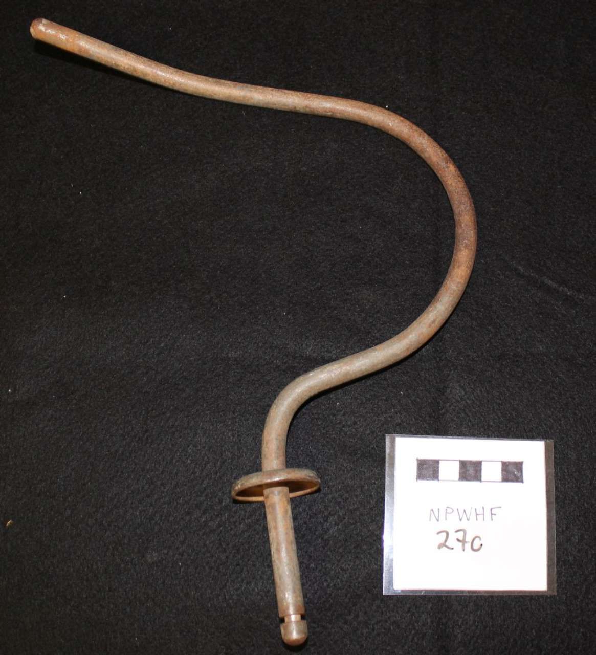 Curved beige metal rod with a ring on black background, beside a small labeled scale card