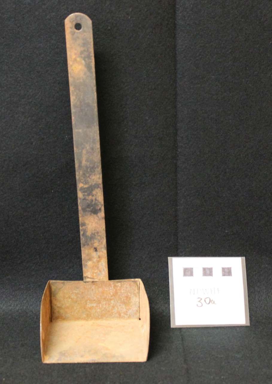 Rusty metal scoop with a long handle standing upright beside a small white label on a dark background