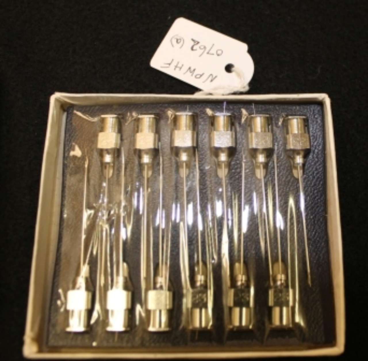 Box of six small glass tubes with metal caps, arranged in a tray, with a handwritten label above them