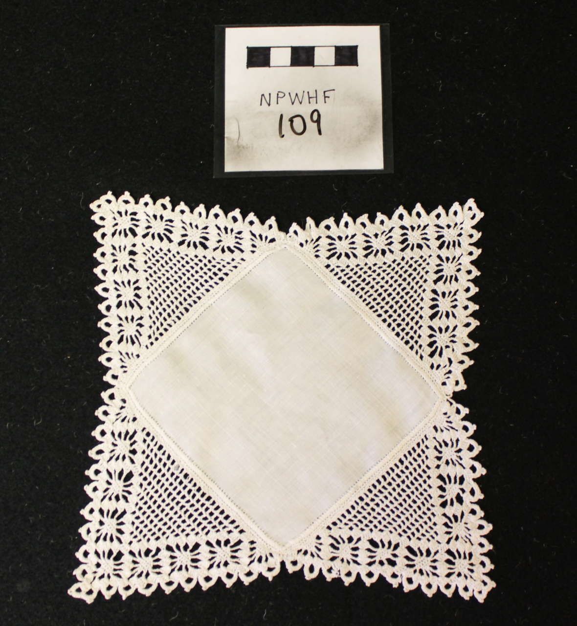 White lace doily with a diamond-shaped center on a black background, with a small labeled sample card above it