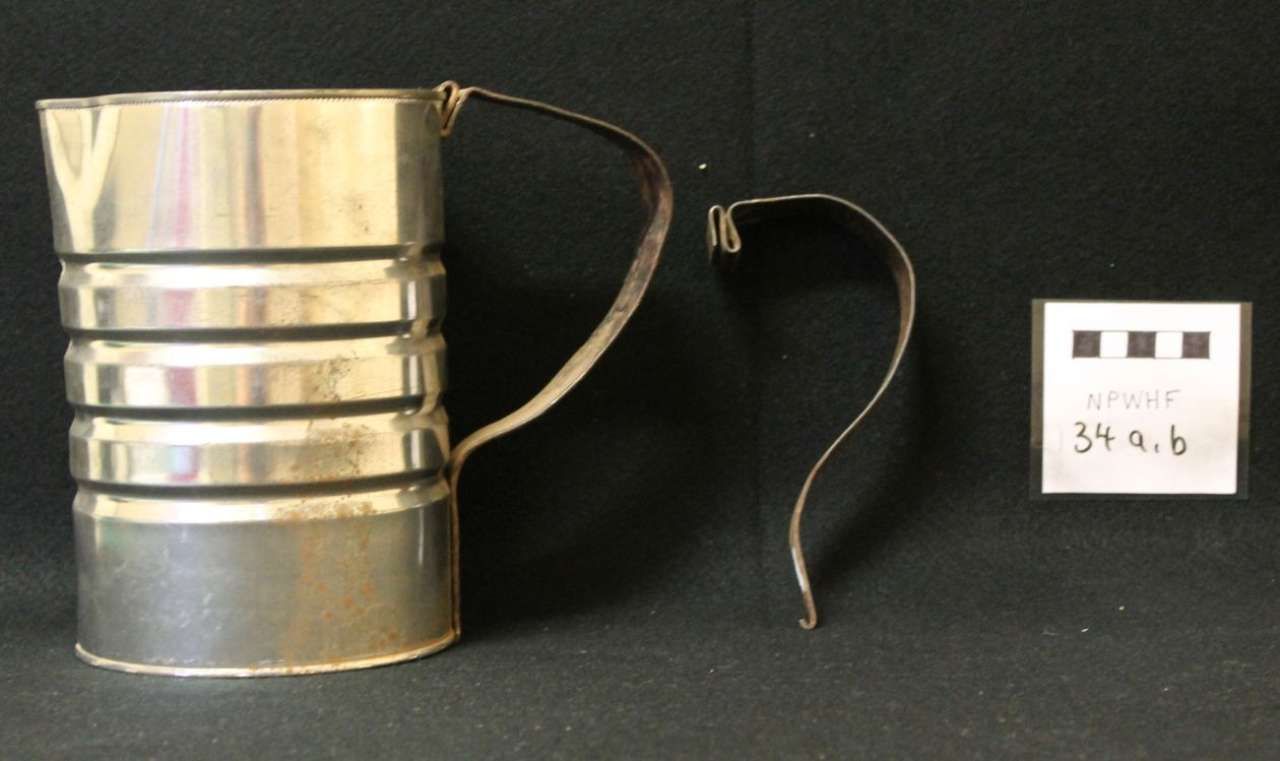 Metal measuring cup with handle and a loose curved wire piece beside a small numbered label on a black background