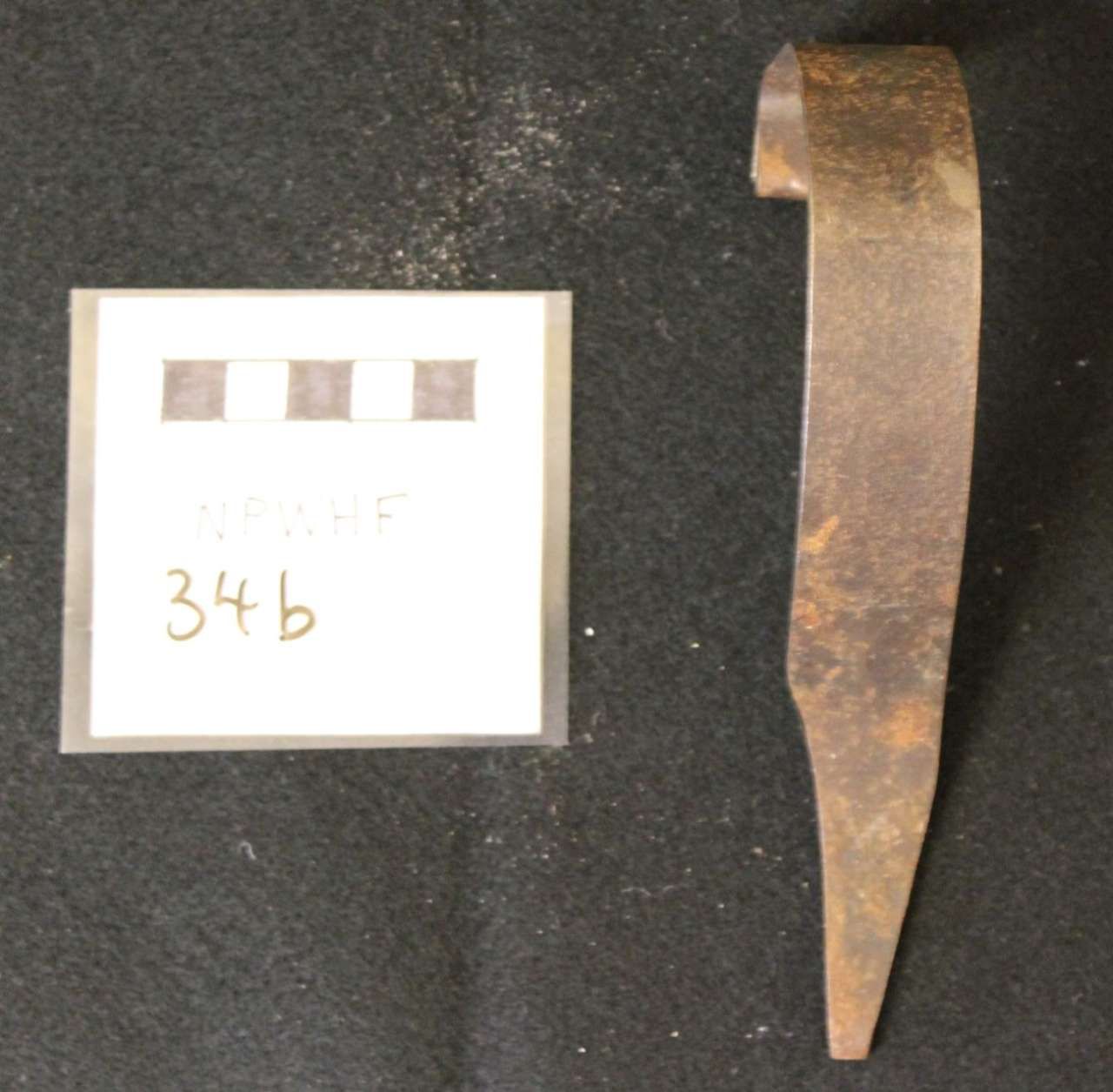 Rusty metal tool or blade fragment beside a labeled white evidence card on a dark background