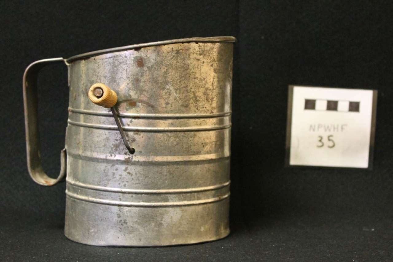 Metal flour sifter with handle and crank on a black background, labeled 