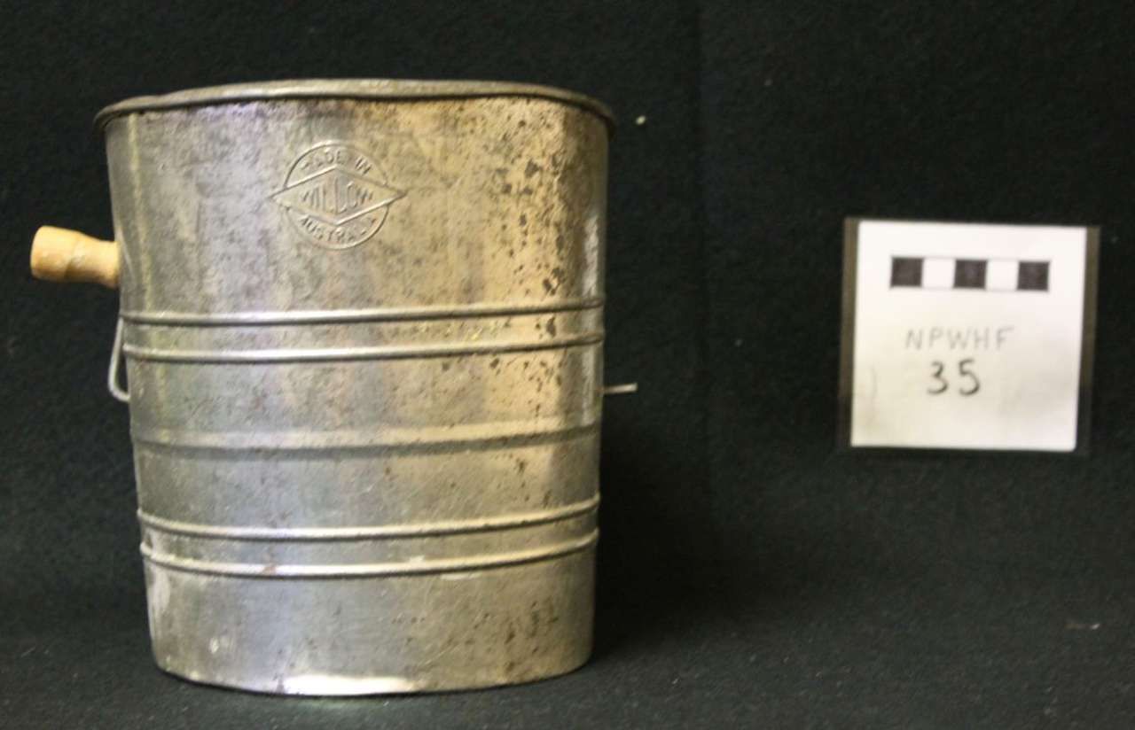 Old metal bucket with a handle and spout against a black background, next to a small label marked “35”