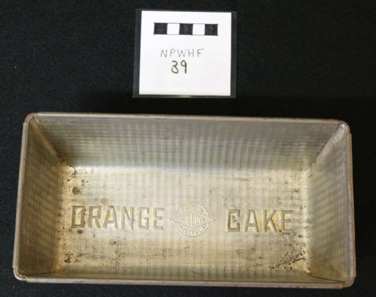 Rectangular metal baking pan with faded orange cake text, labeled tag above on black background