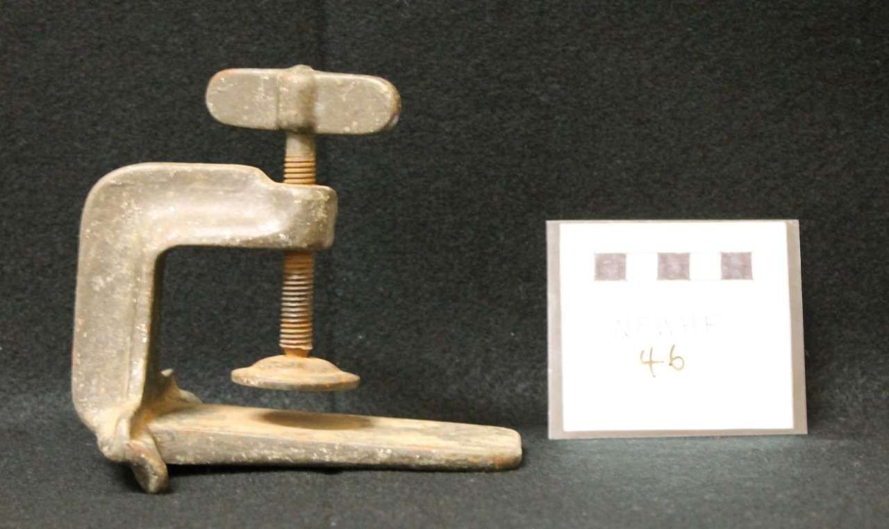 Small metal C-clamp beside a white square card with handwritten “46” on a dark background