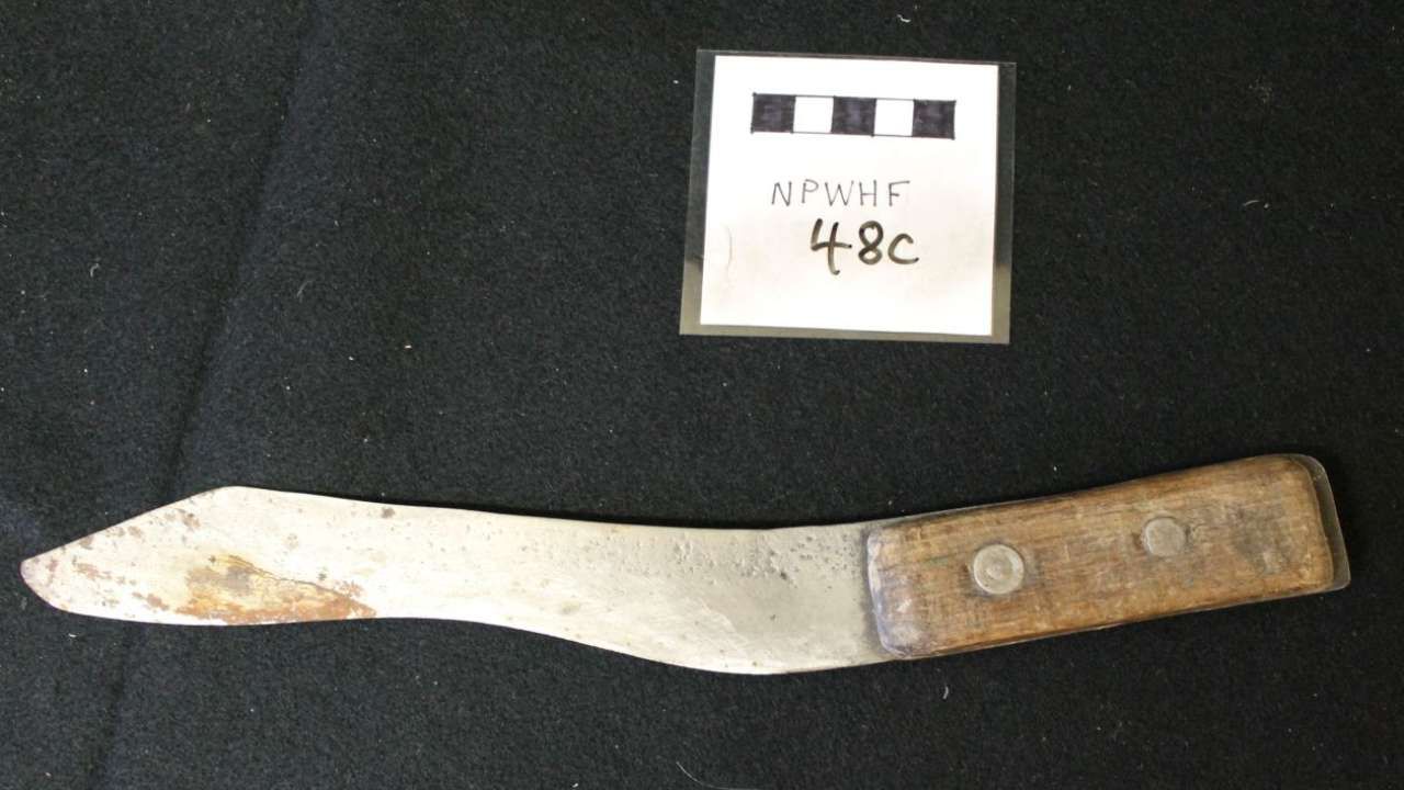 Rusty metal knife with wooden handle laid on a dark surface beside a small labeled tag.