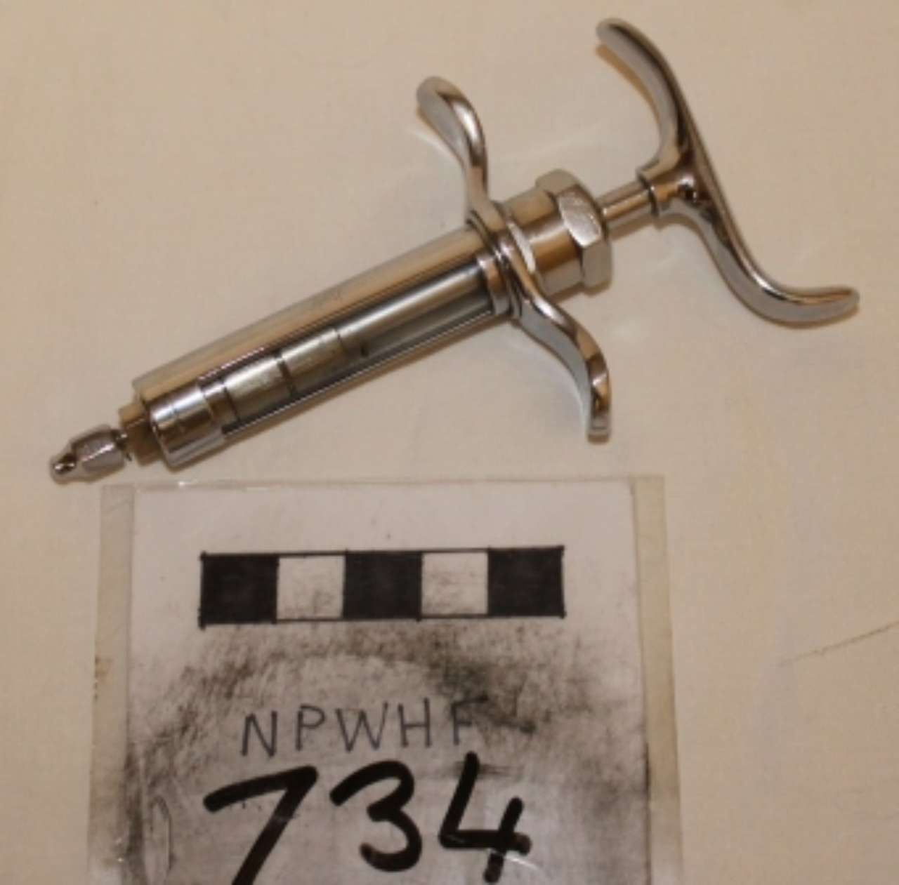 Metal syringe above a labeled evidence card with “734” on a beige background