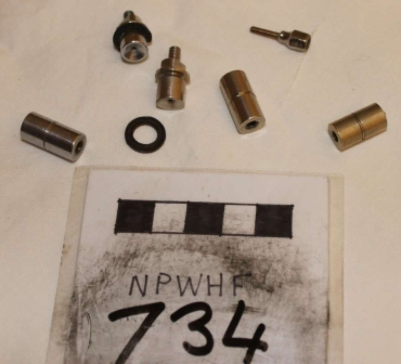 Small metal parts and a black ring arranged above a labeled card reading “NPWFR 734”