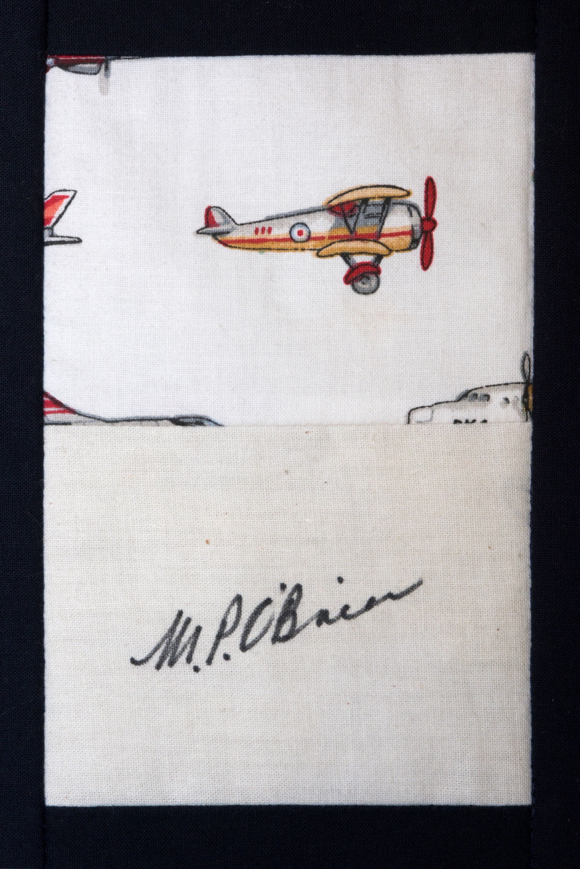 Small embroidered airplane above a black cursive signature on white fabric