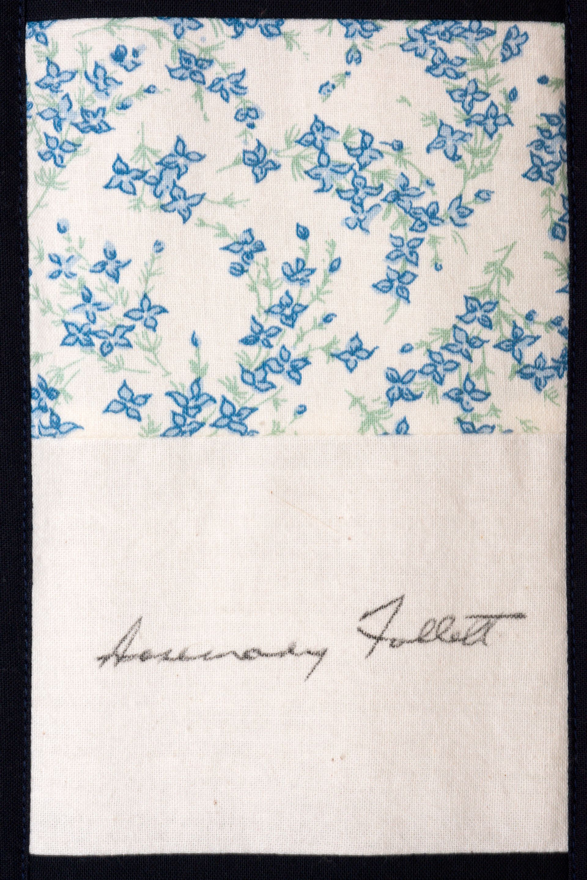 White fabric with blue floral embroidery and handwritten signature near the bottom.