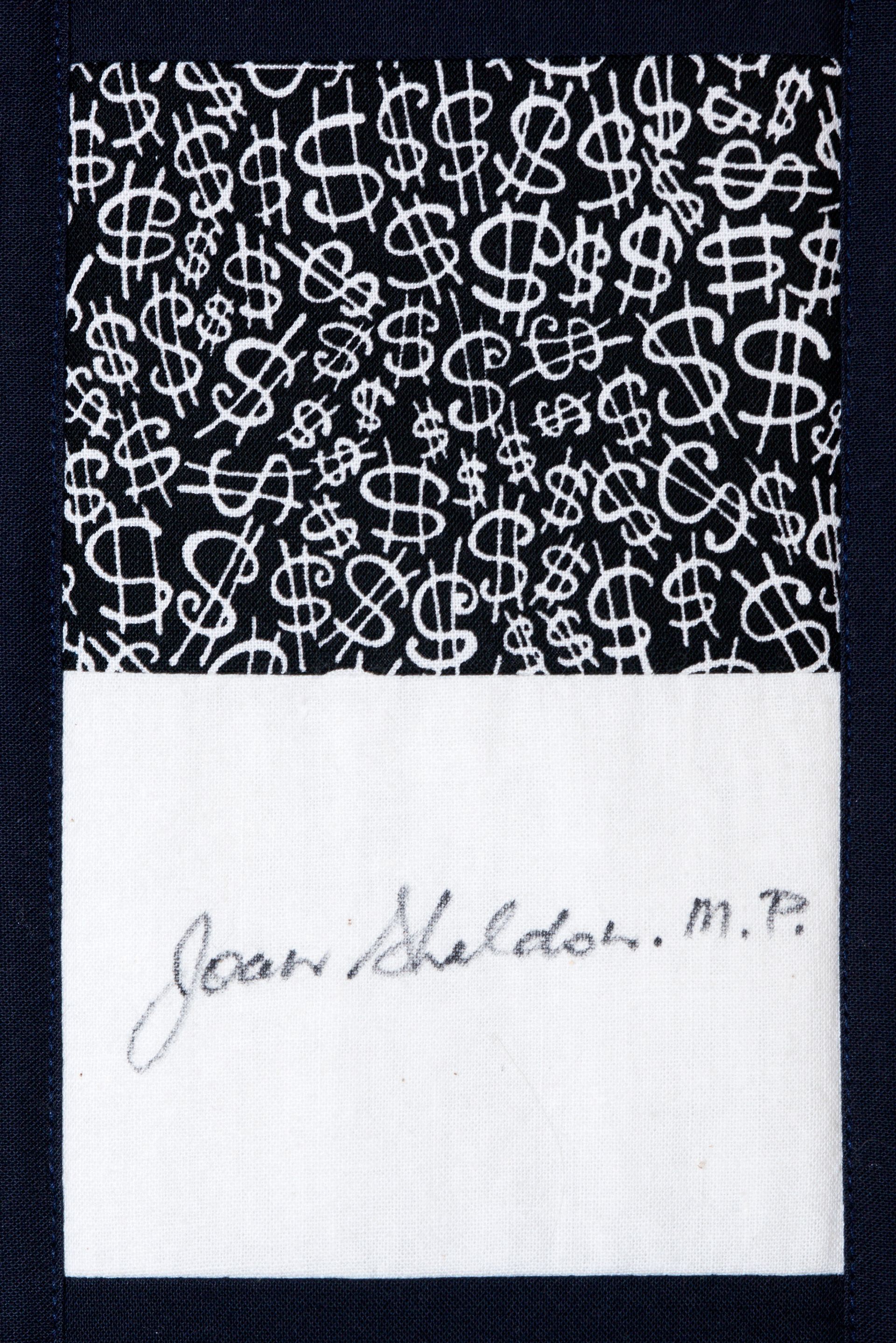 Black fabric with white dollar signs above a white label signed “Jean Sheldon, M.P.”