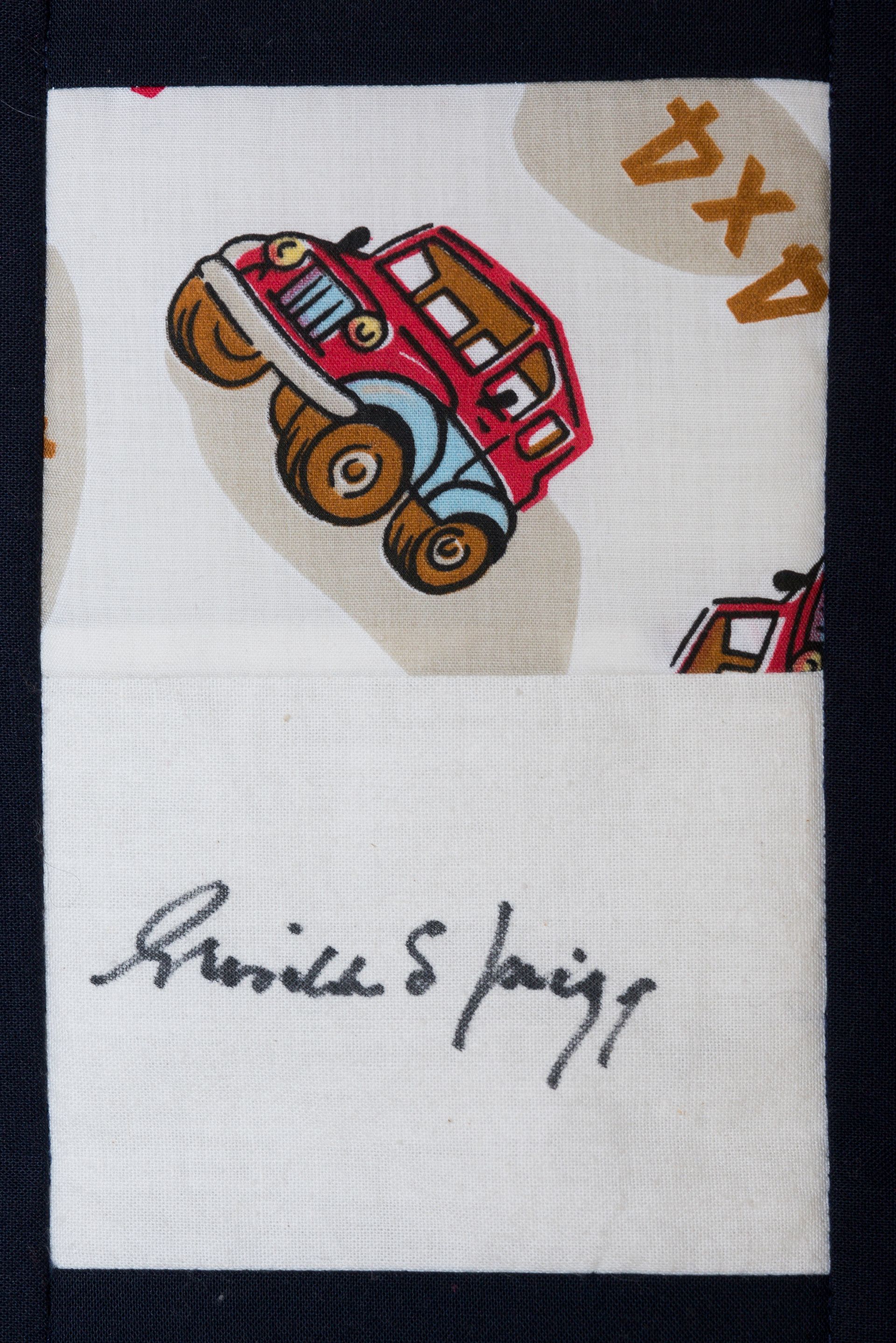 White fabric with a cartoon red car and handwritten signature in black ink, plus small orange marks.