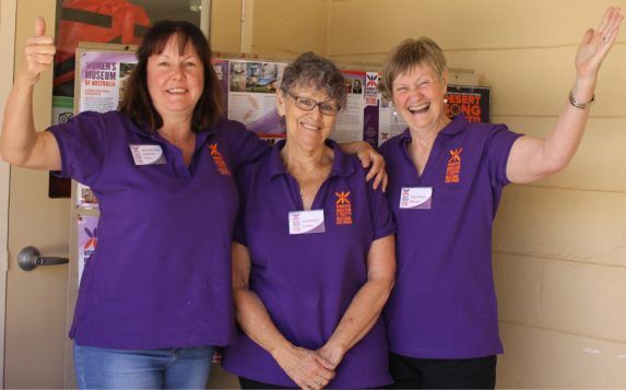 Three people in purple shirts smiling and posing with arms raised indoors