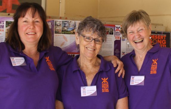 Three women in purple shirts smiling at an indoor event booth.