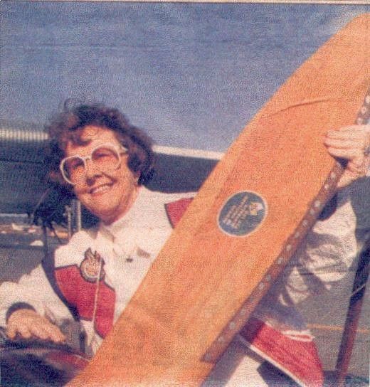 Smiling person holding a large wooden paddle with a circular emblem outdoors under a blue sky